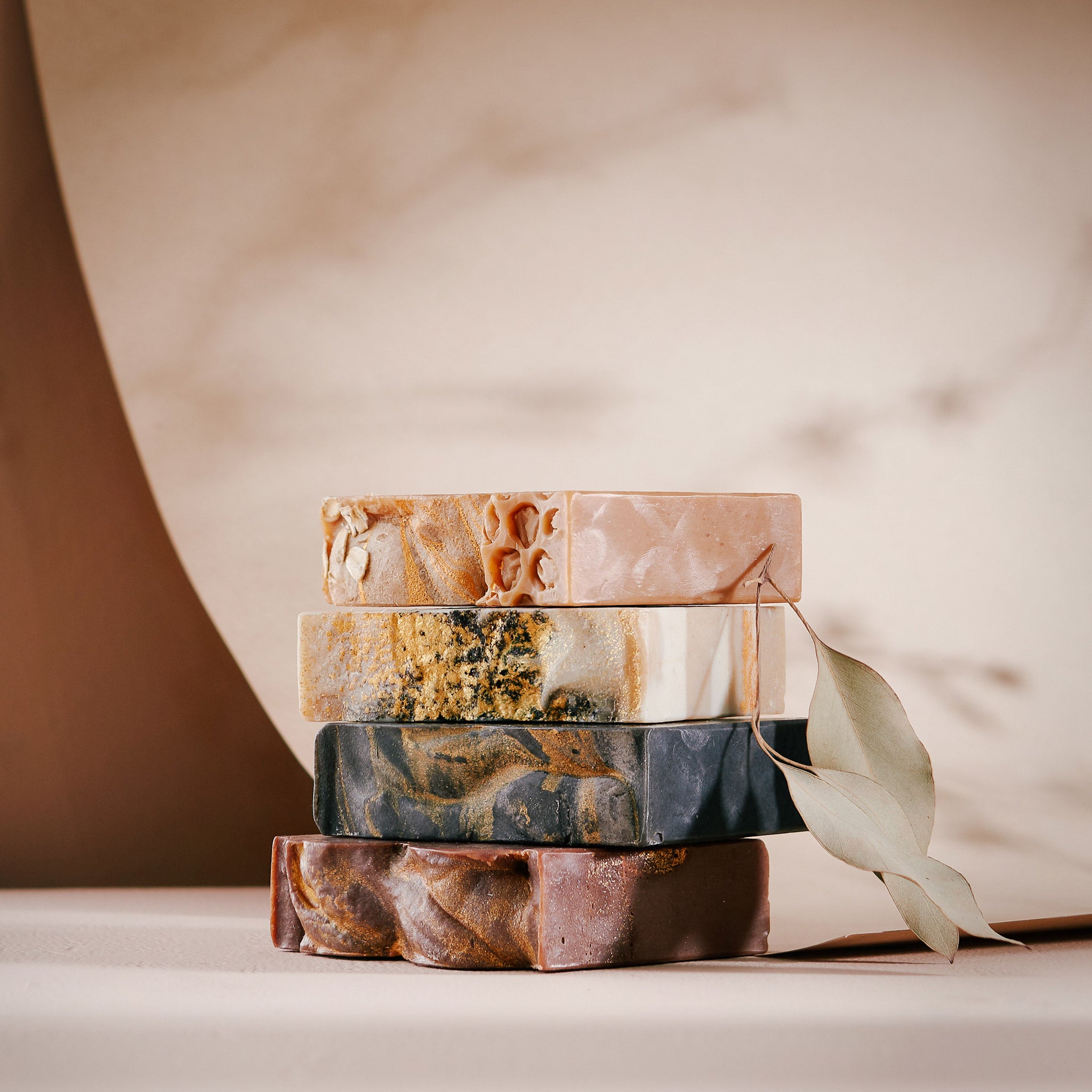 A range of Sonture's handmade soaps sit stacked in a lightly shadowed beige background. The soaps feature warm colours and gold swirls.