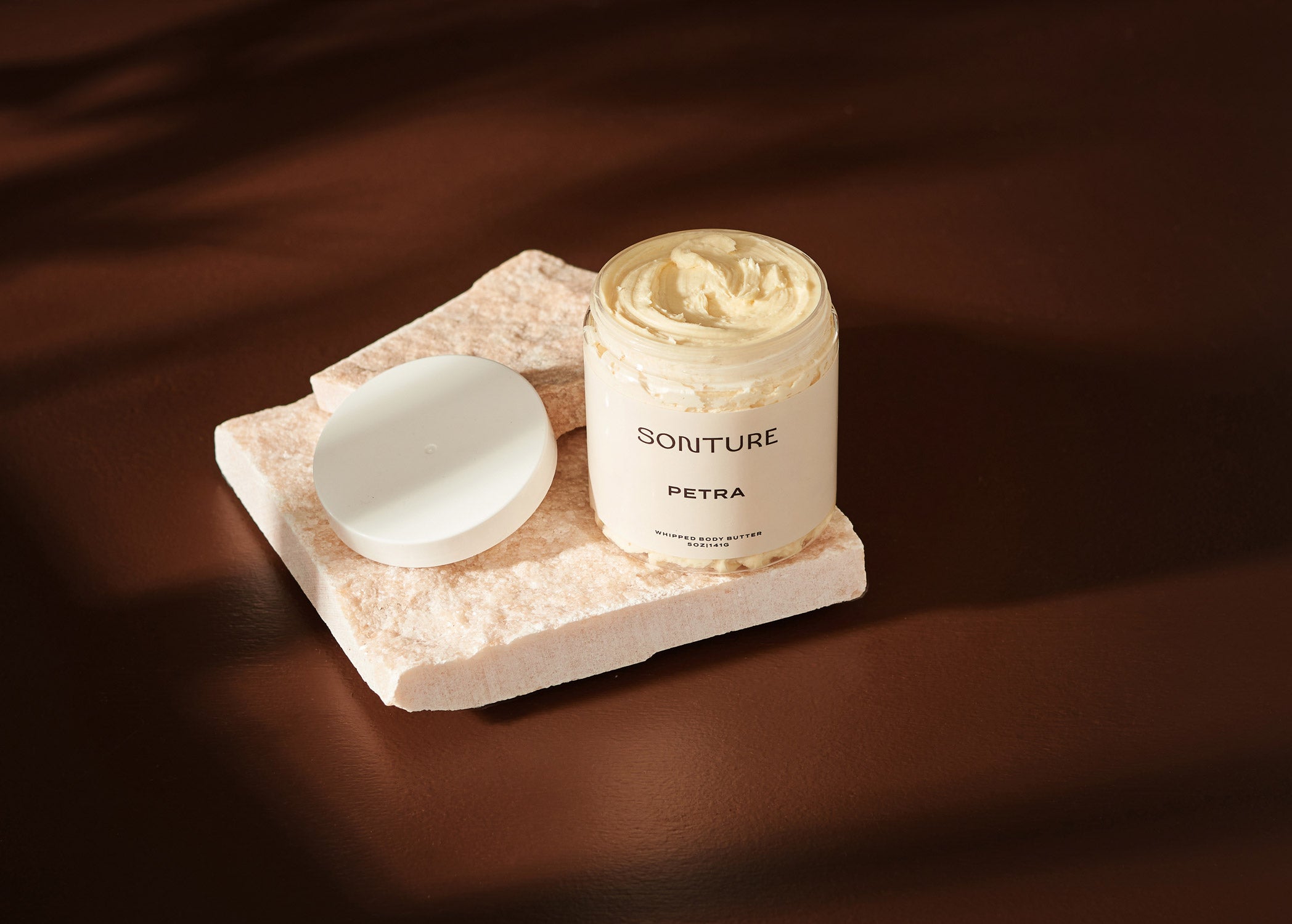 Sonture handmade Petra body butter sits on a mineral tile in a deep brown shadowy setting. The body butter container is clear (so as to show the detail of the product) and has a luxurious whipped texture.