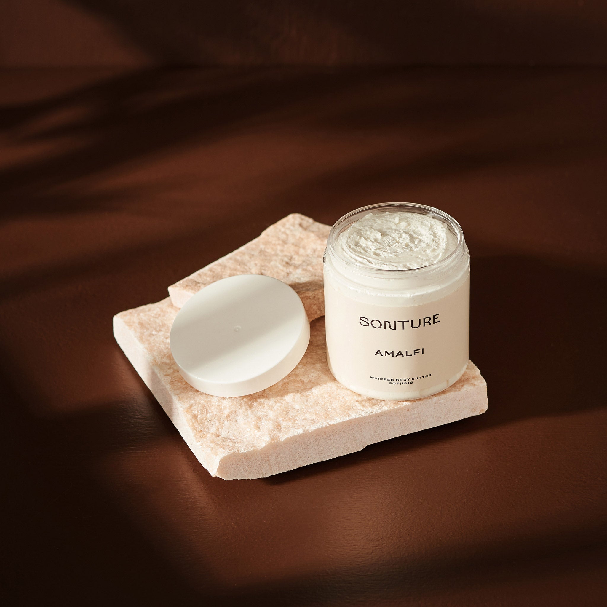 Sonture handmade Amalfi body butter sits on a mineral tile in a deep brown shadowy setting. The body butter container is clear (so as to show the detail of the product) and has a luxurious whipped texture.