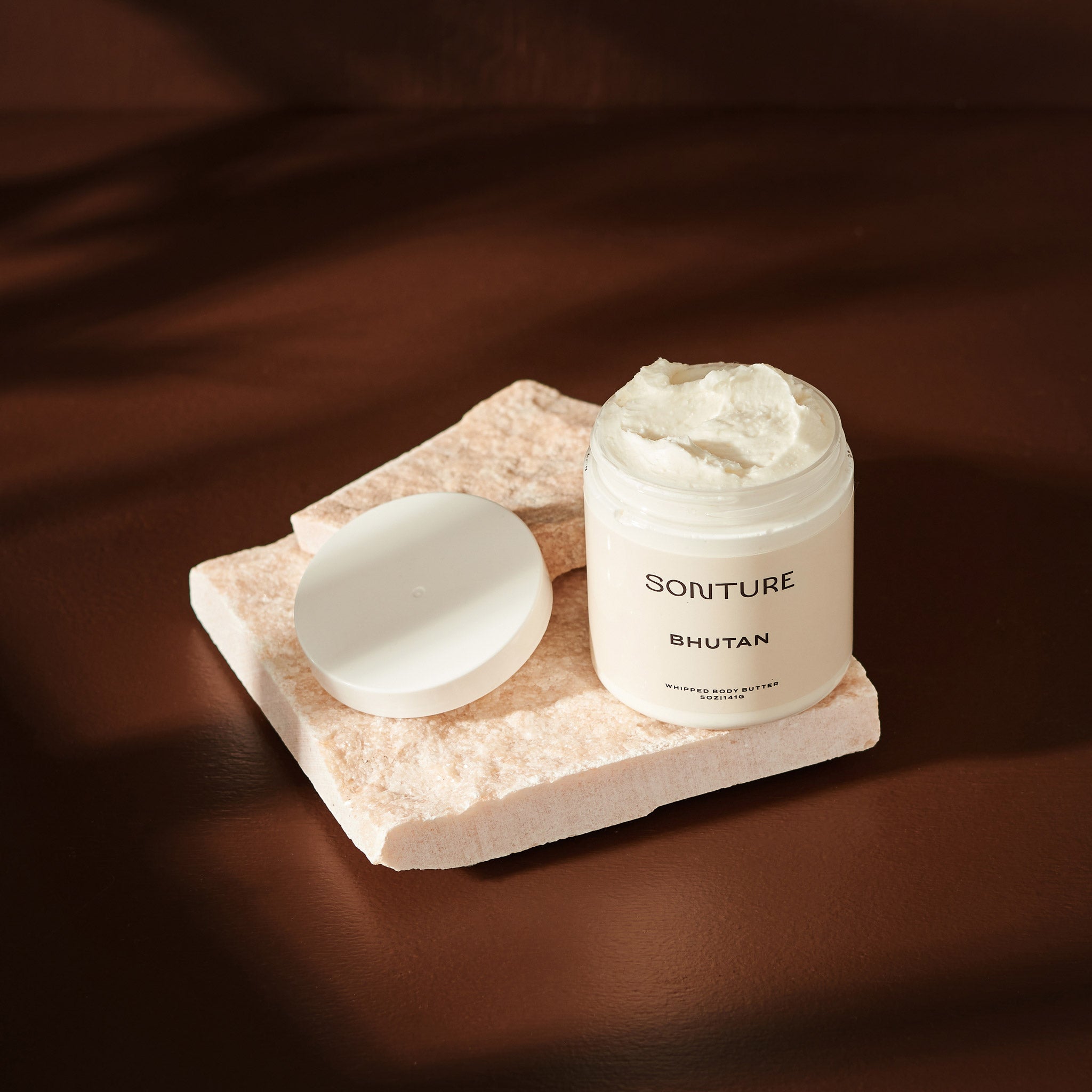 Sonture handmade Bhutan body butter sits on a mineral tile in a deep brown shadowy setting. The body butter container is clear (so as to show the detail of the product) and has a luxurious whipped texture.