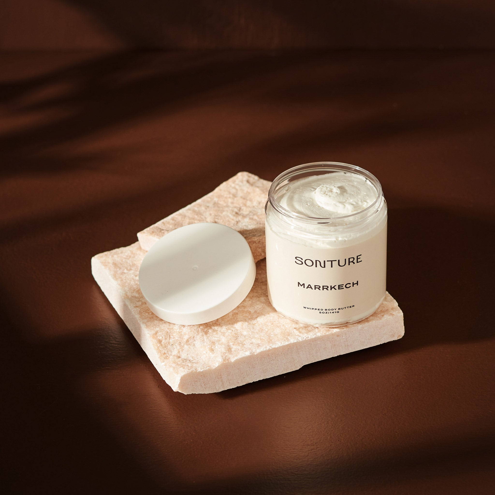 Sonture handmade Marrakech body butter sits on a mineral tile in a deep brown shadowy setting. The body butter container is clear (so as to show the detail of the product) and has a luxurious whipped texture.