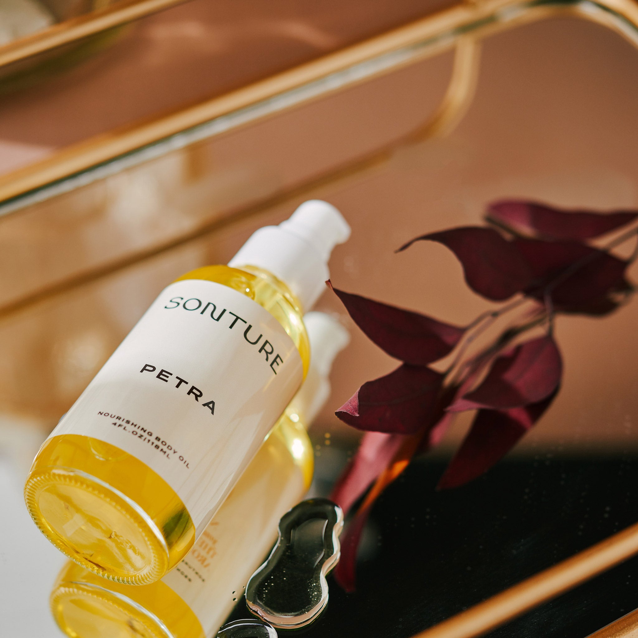 Sonture’s Petra Nourishing handcrafted body oil is travel inspired and features ethical ingredients. Here the oil sits on a gold tray with a deep purple leaf, the oil dripped on the mirrored surface.