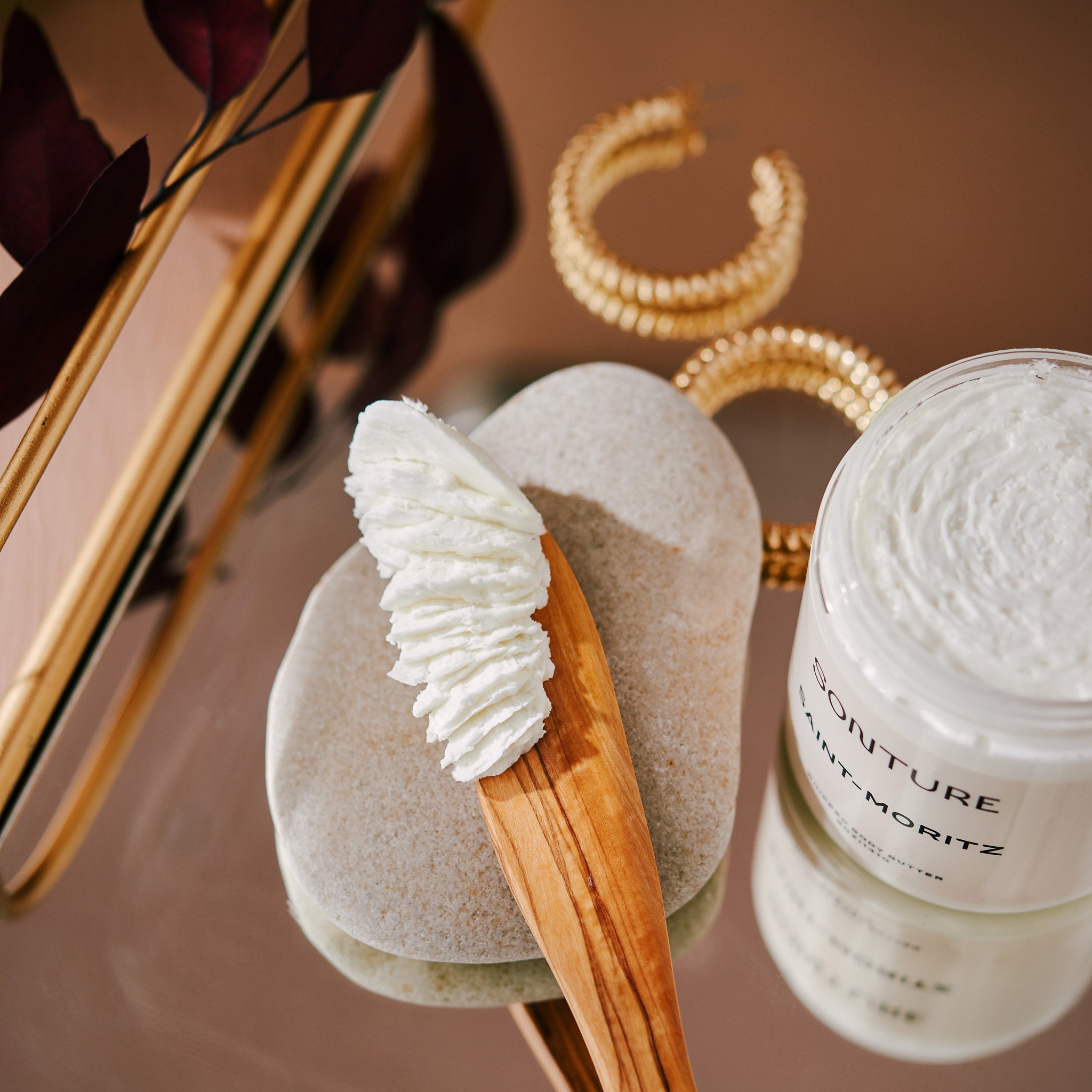 Sonture's Saint-Moritz whipped body butter is styled on a wooden scoop on a glamorous gold mirrored tray.