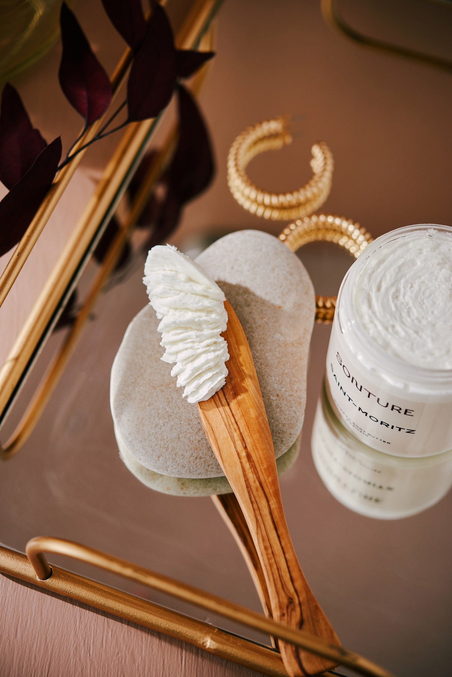 The Sonture handmade whipped body butter Saint-Motitz is styled on an elegant gold tray with gold earrings and botanical elements. The travel-inspired bath product is in a clear container with a white lid and cream coloured label.