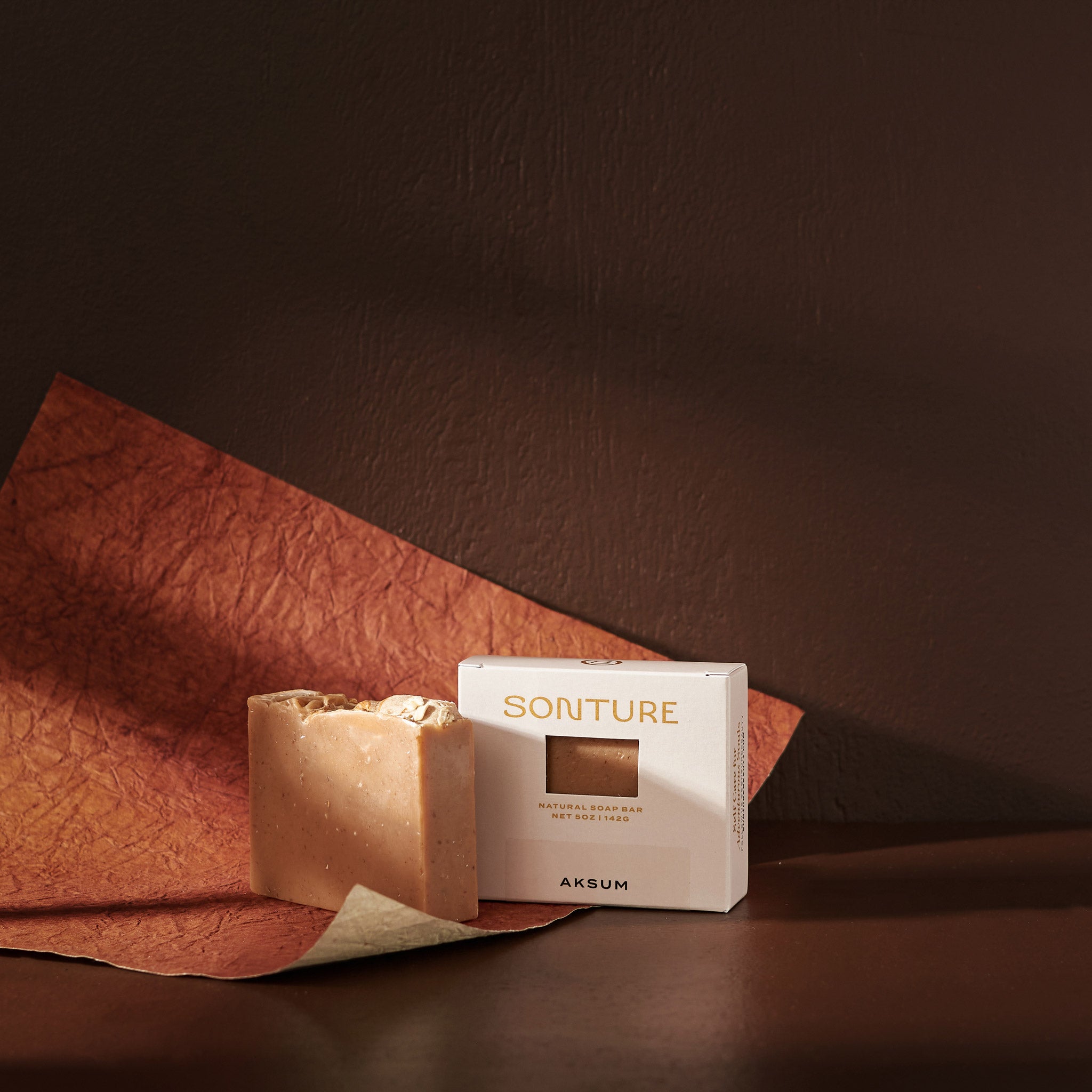 Sonture’s Aksum handcrafted natural soap bar is inspired by travel and features nourishing natural ingredients. Here the luxurious self care product sits in a cream coloured box with a diecut in a deep chocolate brown background with rust red paper. 