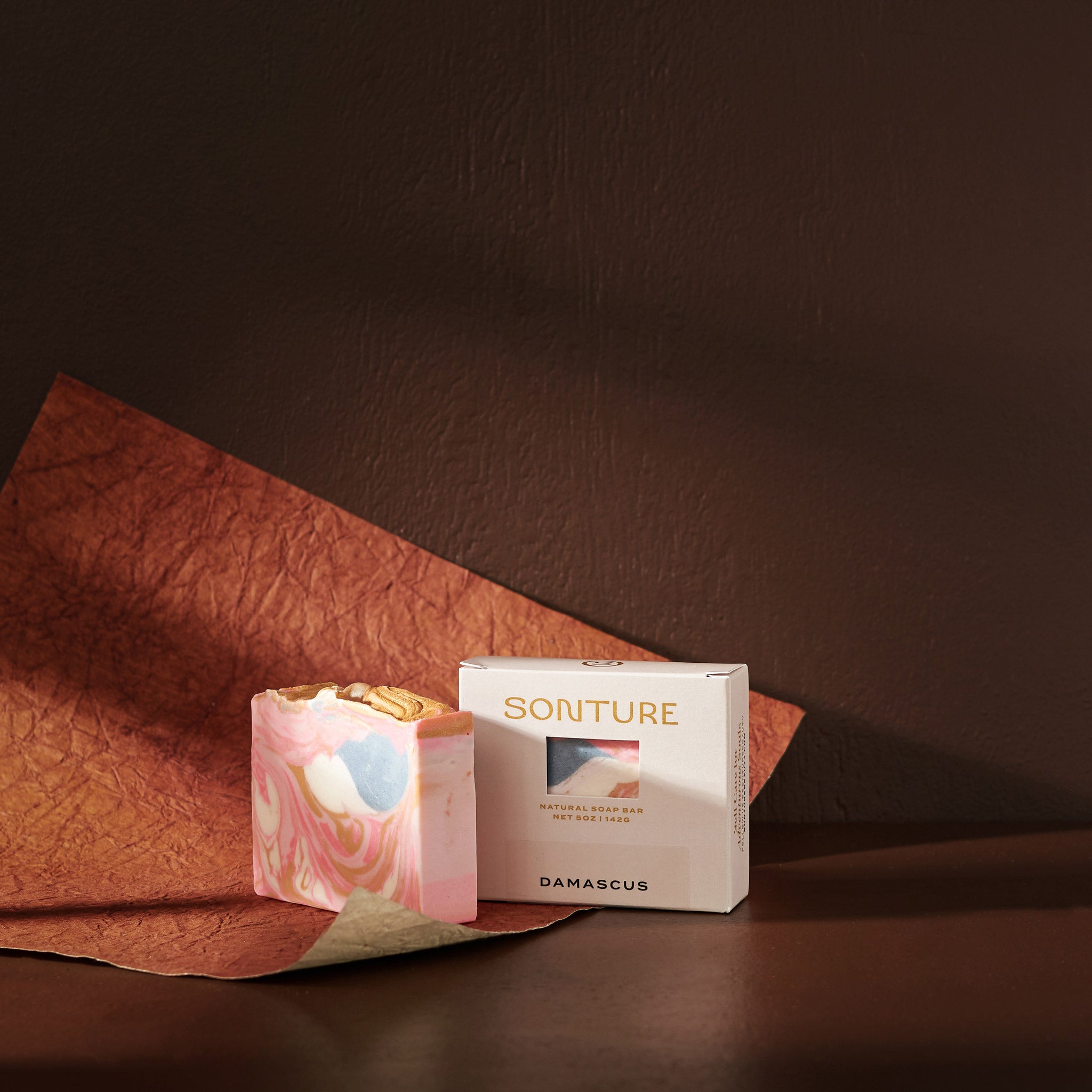 Sonture’s Damascus handcrafted natural soap bar is inspired by travel and features nourishing natural ingredients. Here the luxurious self care product sits in a cream coloured box with a diet in a deep chocolate brown background with rust red paper. 