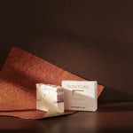 Sonture’s Marseille handcrafted natural soap bar is inspired by travel and features nourishing natural ingredients. Here the luxurious self care product sits in a cream coloured box with a diet in a deep chocolate brown background with rust red paper. 