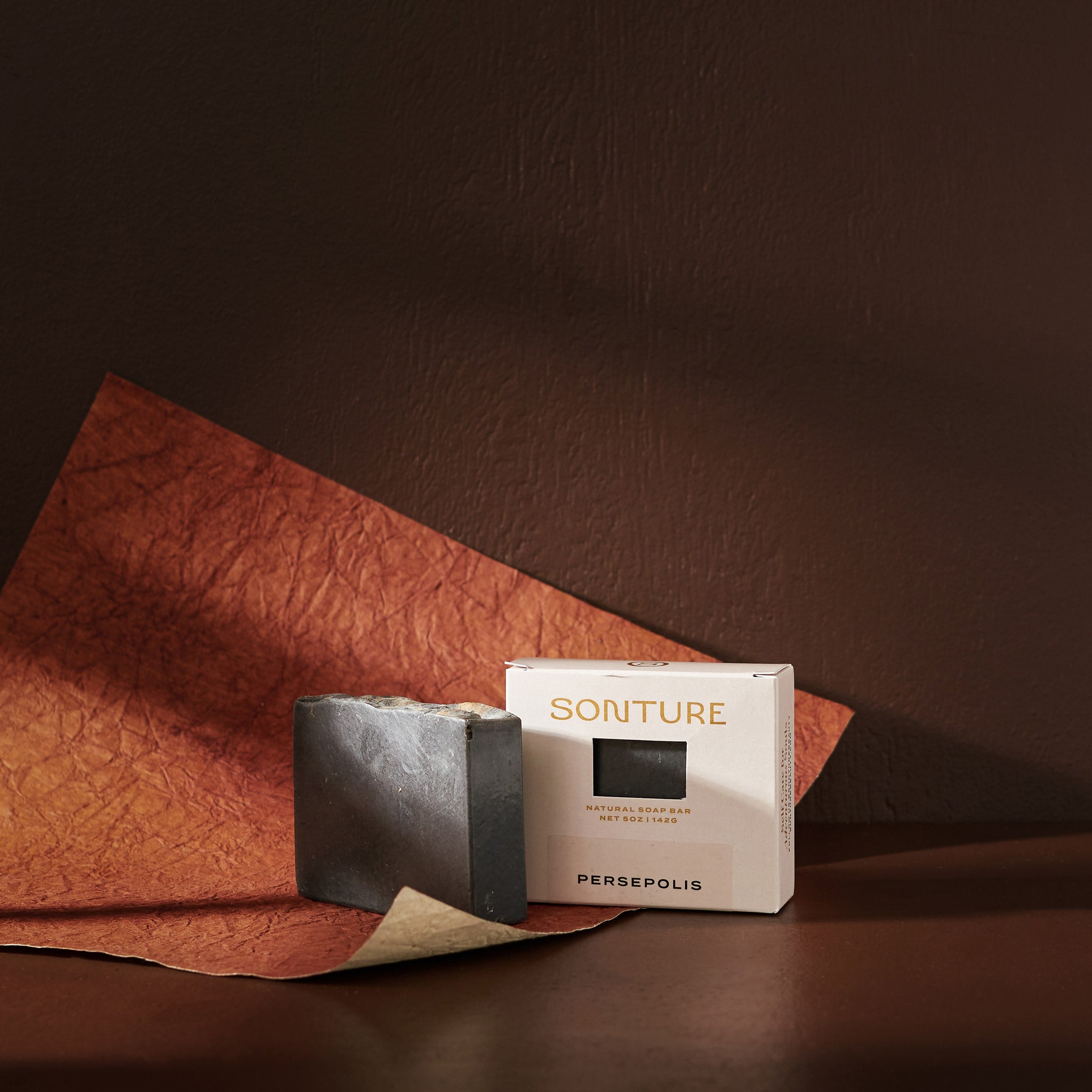 Sonture’s Persepolis handcrafted natural soap bar is inspired by travel and features nourishing natural ingredients. Here the luxurious self care product sits in a cream coloured box with a diet in a deep chocolate brown background with rust red paper. 